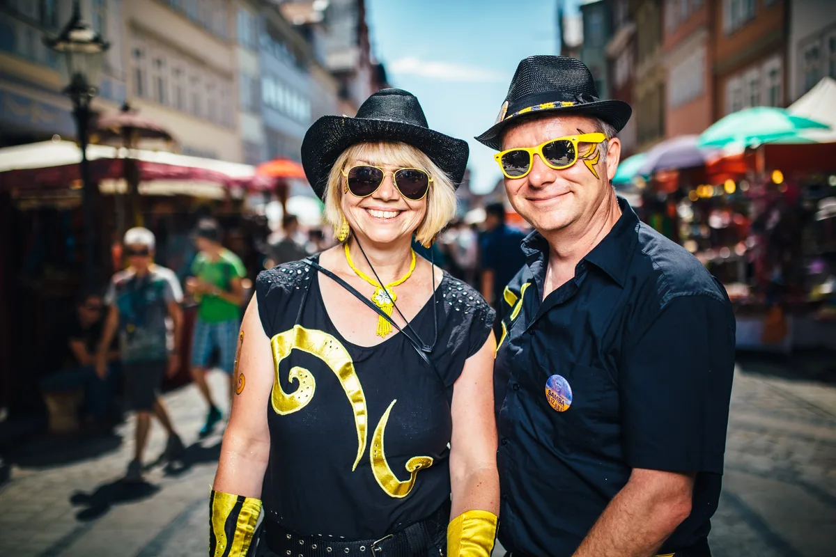 Dasselbe Paar enger im Bild, links Frau mit gelber Kette, rechts Mann mit gelber Sonnenbrille und Gesichtsmalerei