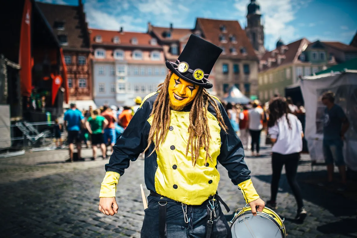 Trommler mit gelb geschminktem Gesicht, Zylinder mit Schutzbrille und Dreadlocks hält lächelnd seine Trommel