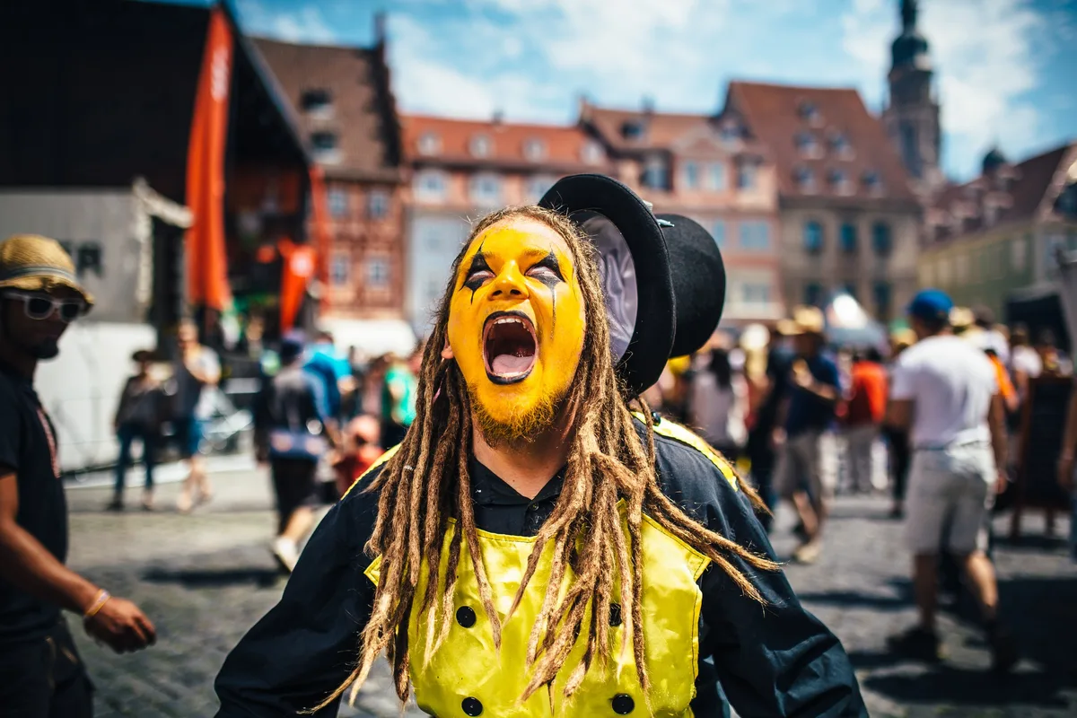 Gelb geschminkter Trommler mit Dreadlocks und Zylinder schreit mit weit offenem Mund in die Kamera