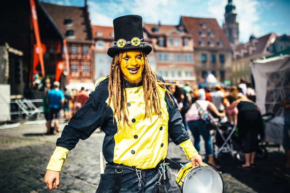 Trommler mit gelber Gesichtsbemalung lacht mit offenem Mund, Zylinder mit Biohazard-Brille, Altstadthäuser dahinter