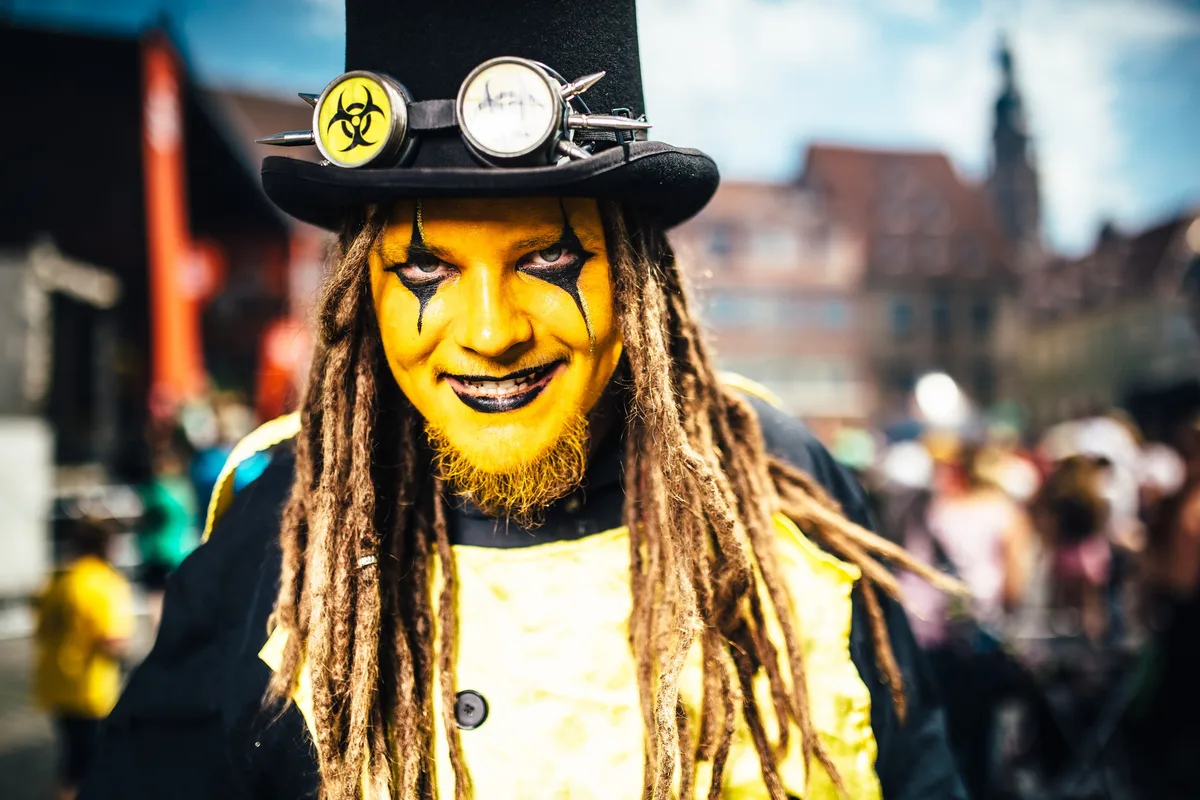 Nahaufnahme des gelb geschminkten Trommlers mit schwarzen Augenzeichnungen, Zylinder mit Stachelbrille und Dreadlocks