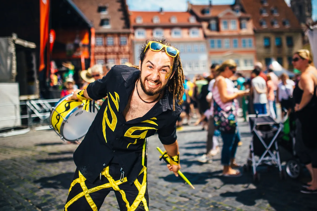 Trommler mit Dreadlocks und gelber Sonnenbrille im Haar grinst breit und beugt sich mit Trommel nach vorn