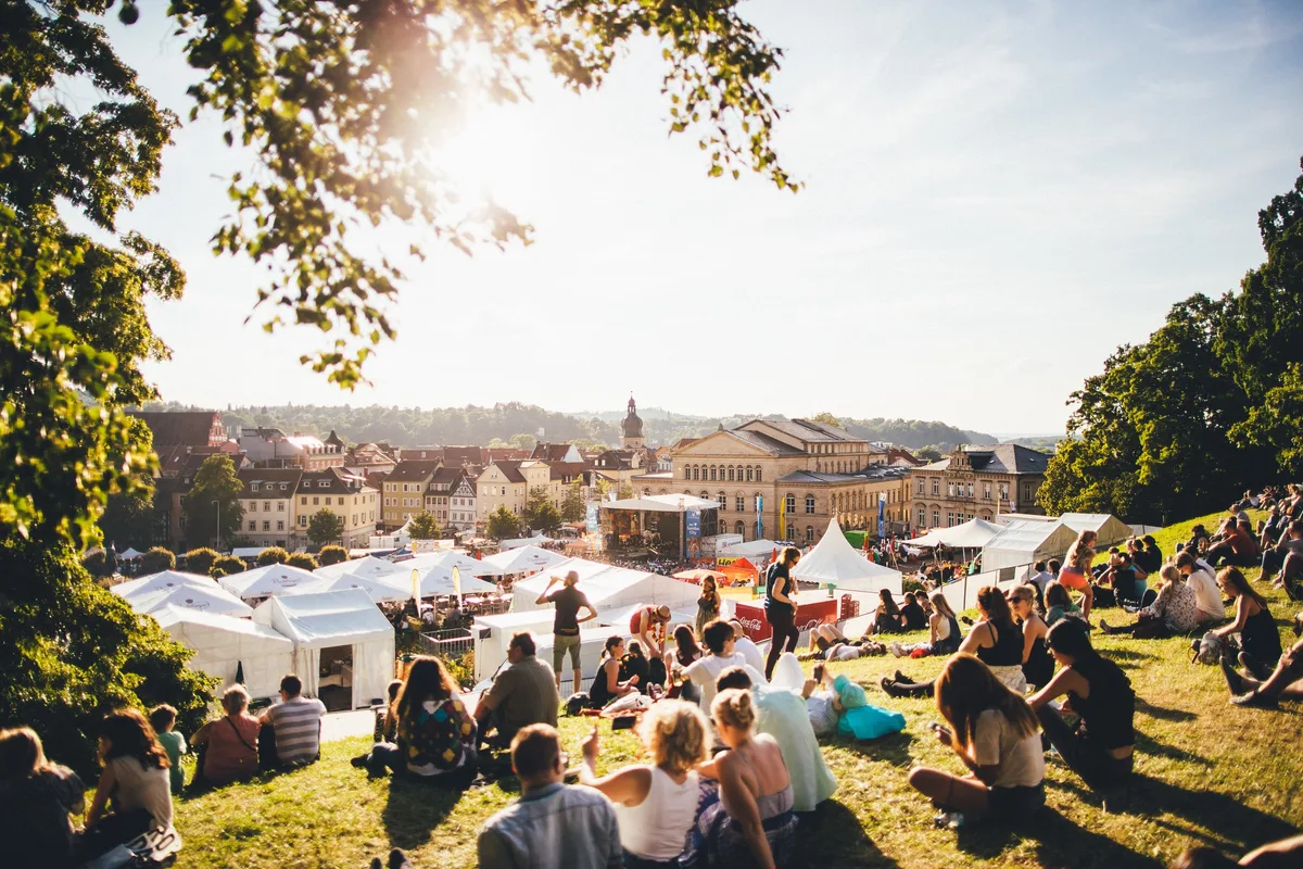 Festivalgäste sitzen im Gegenlicht auf einer Wiese oberhalb der Stadt, unten weiße Zelte und die Bühne