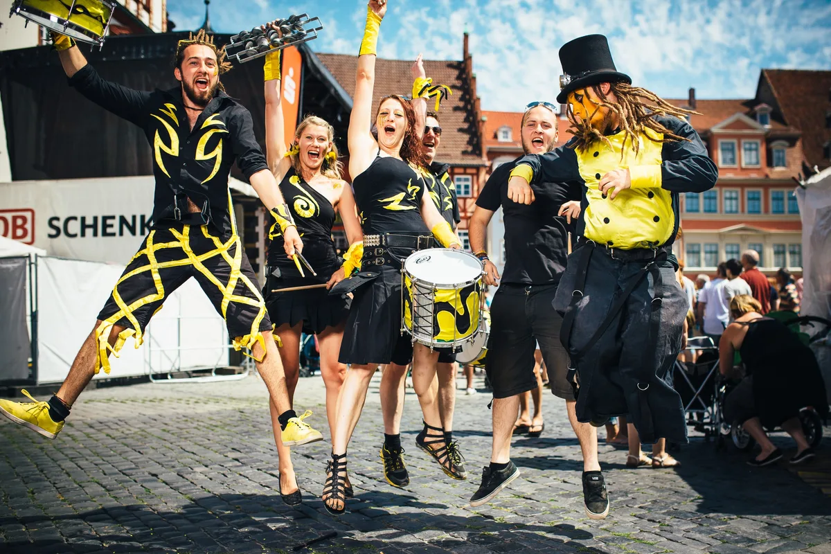 Gruppe von Trommlern und Tänzerinnen in Schwarz und Gelb springt gemeinsam in die Luft, vor Bühne und Altstadthäusern