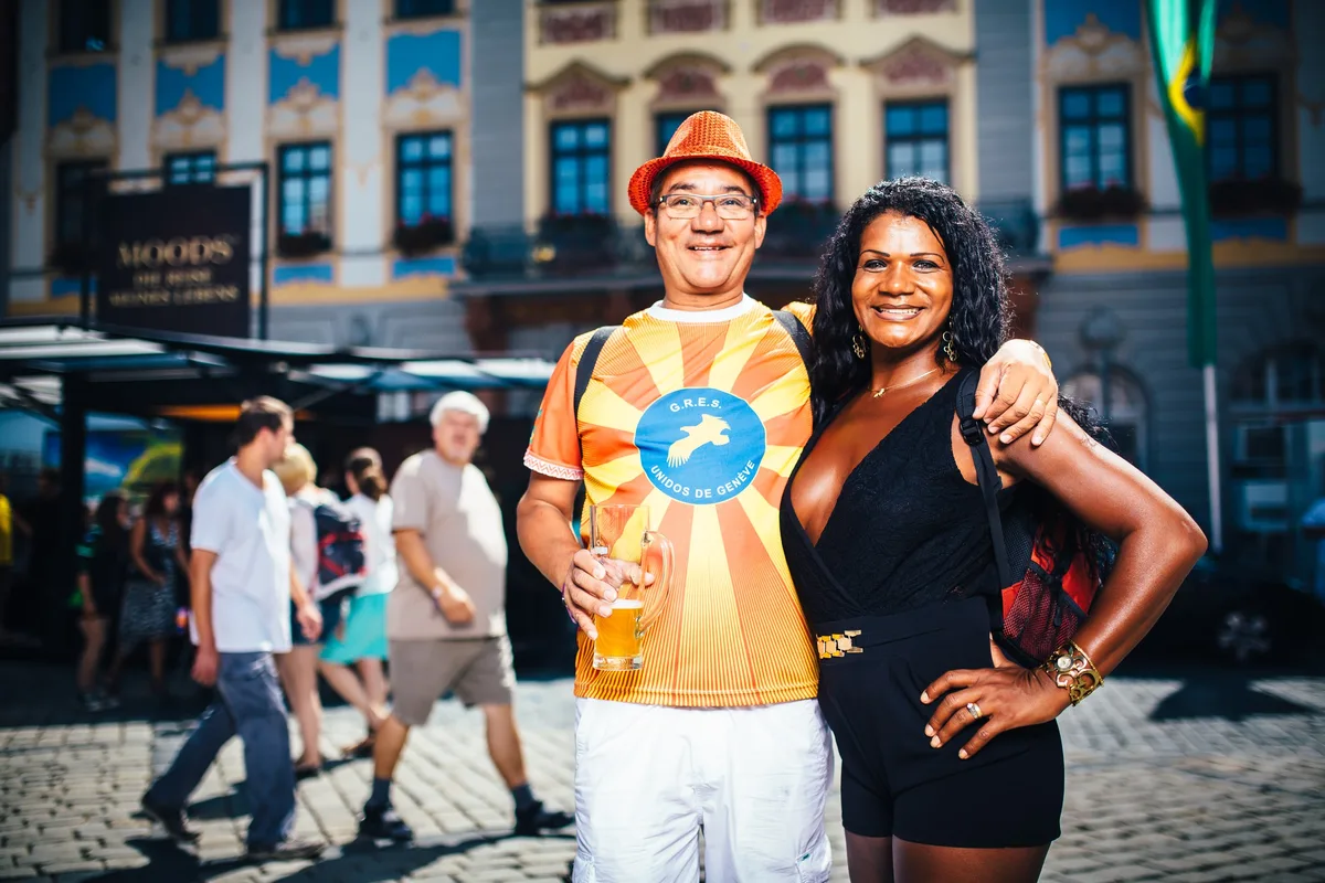 Mann im orangen Hut mit Bierglas und Person mit schwarzen Locken legen sich den Arm um die Schulter, Altstadtplatz dahinter