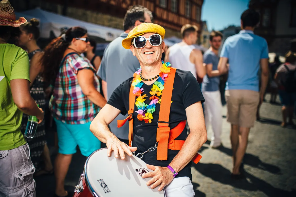 Lachender Trommler mit gelber Mütze, Sonnenbrille und bunter Blumenkette, die Hände auf dem Trommelfell