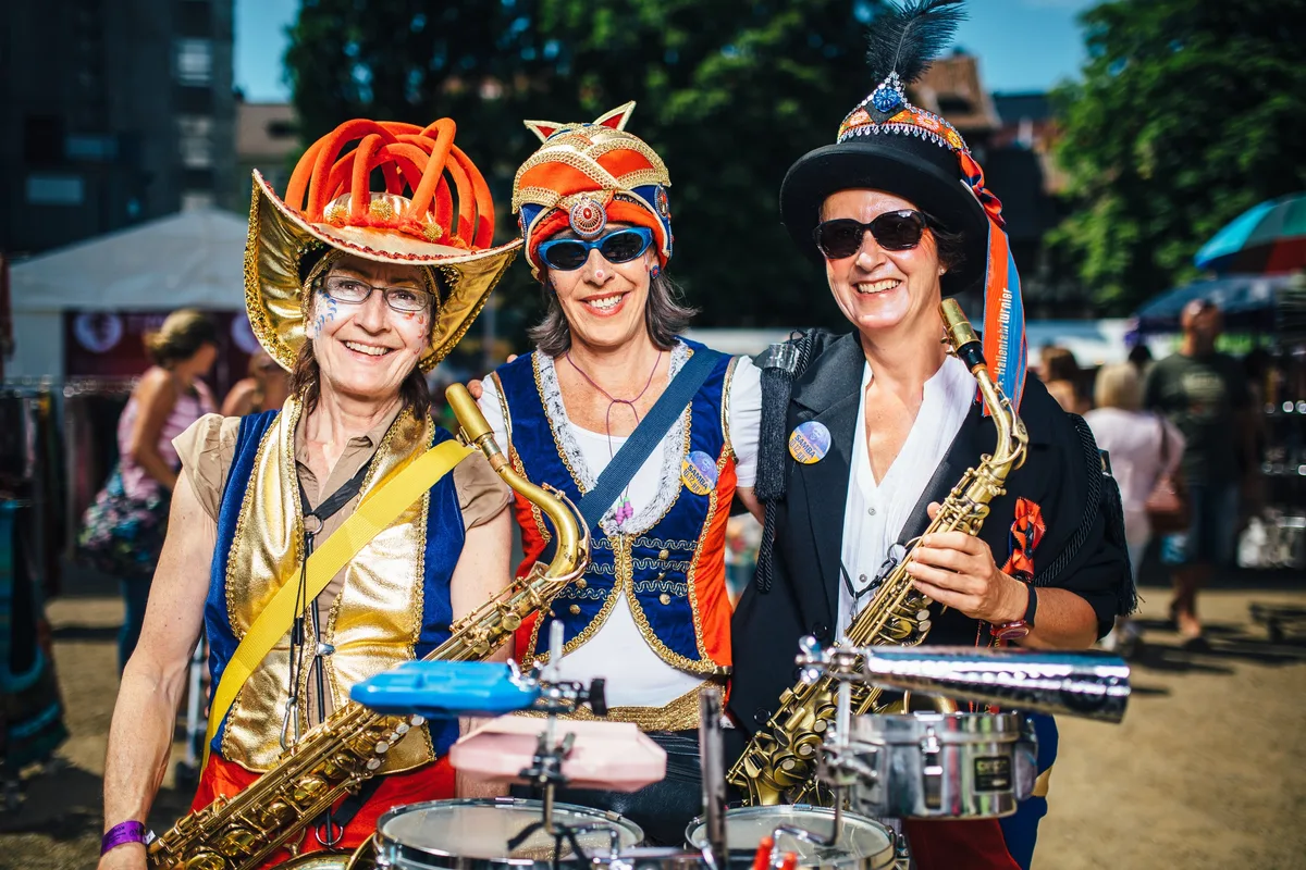 Drei lächelnde Frauen mit Saxofonen in prunkvollen Hüten und Westen, vor ihnen ein kleines Schlagzeug