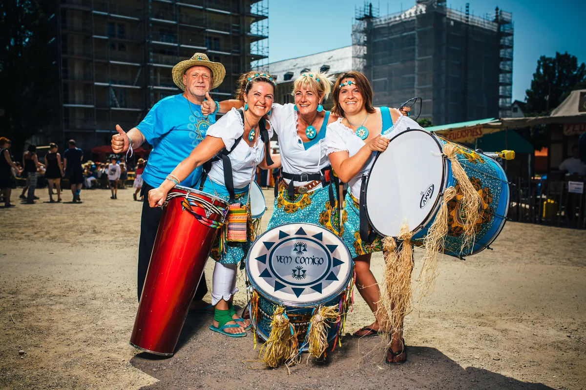 Vier Trommler in Türkis mit Basstrommeln und Conga posieren auf Kiesboden, Gerüst an einem Gebäude im Hintergrund