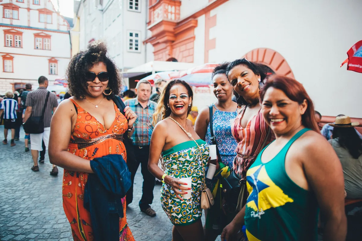 Fünf lachende Frauen in bunten Kleidern und grünem Brasilien-Trikot posieren in einer Gasse, in den Händen Becher