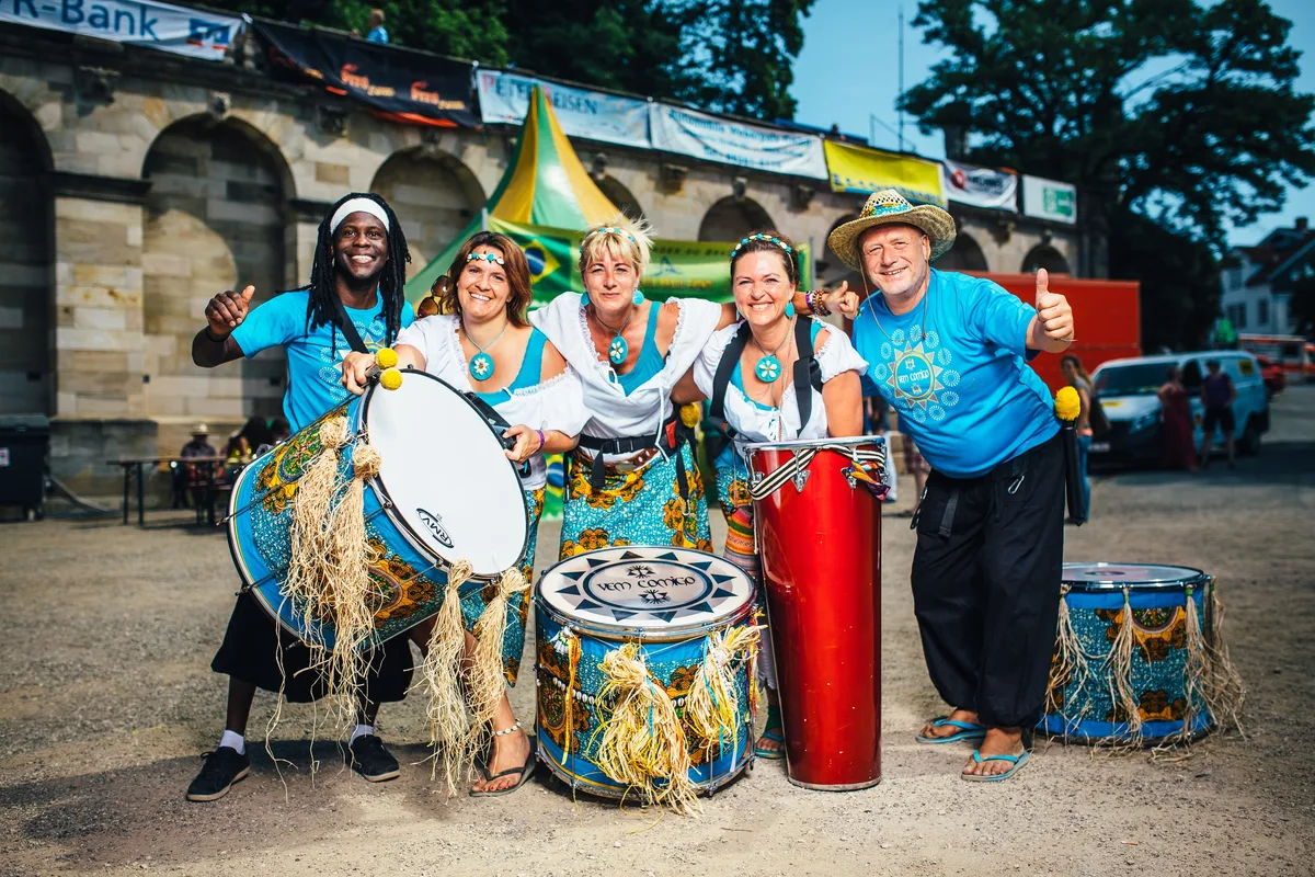 Trommlergruppe in Türkis mit Basstrommeln und roter Conga auf Kiesplatz, beide Männer außen recken die Daumen