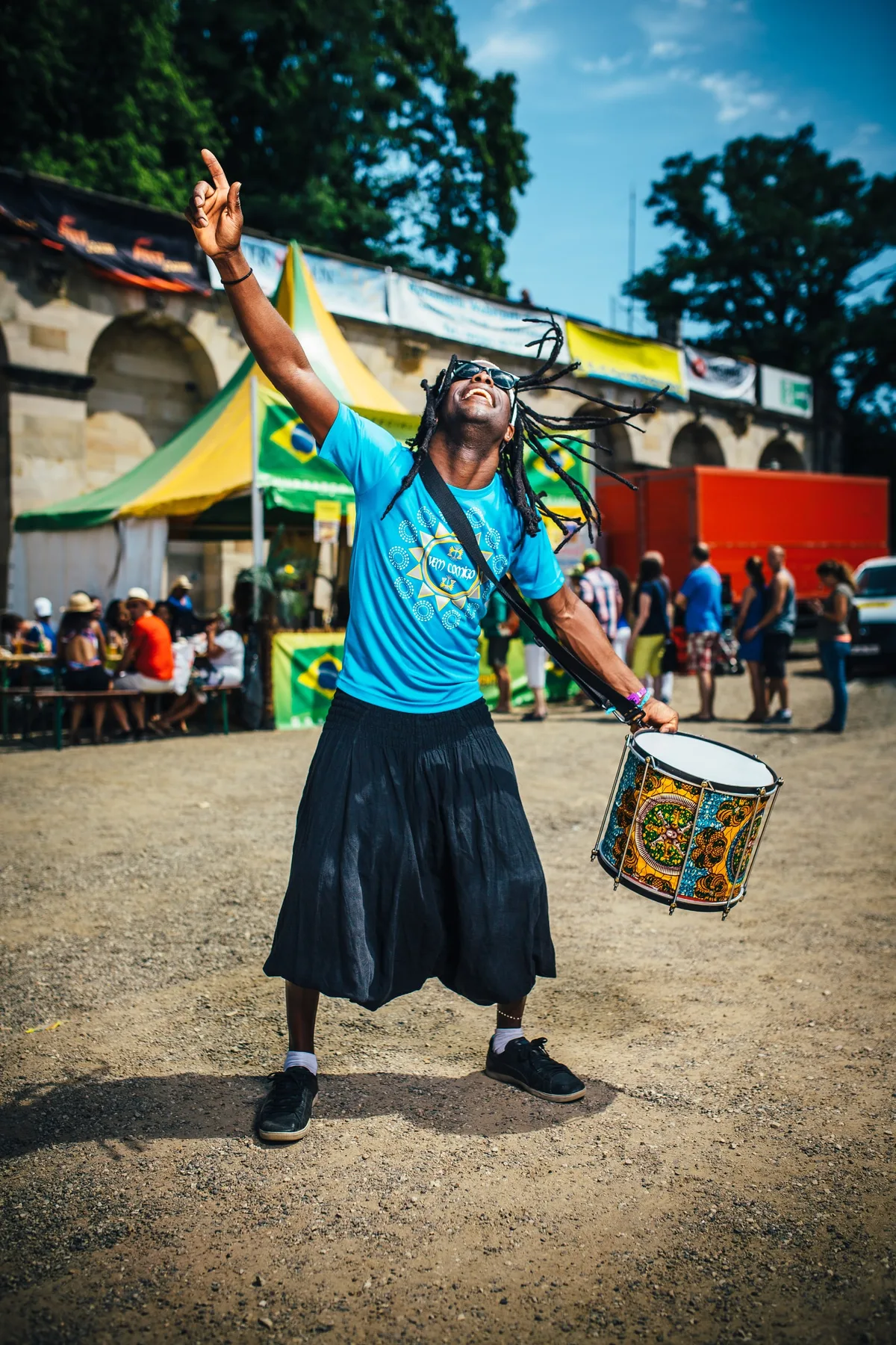 Trommler mit langen Dreadlocks, Sonnenbrille und Umhängetrommel zeigt jubelnd mit einem Arm zum Himmel