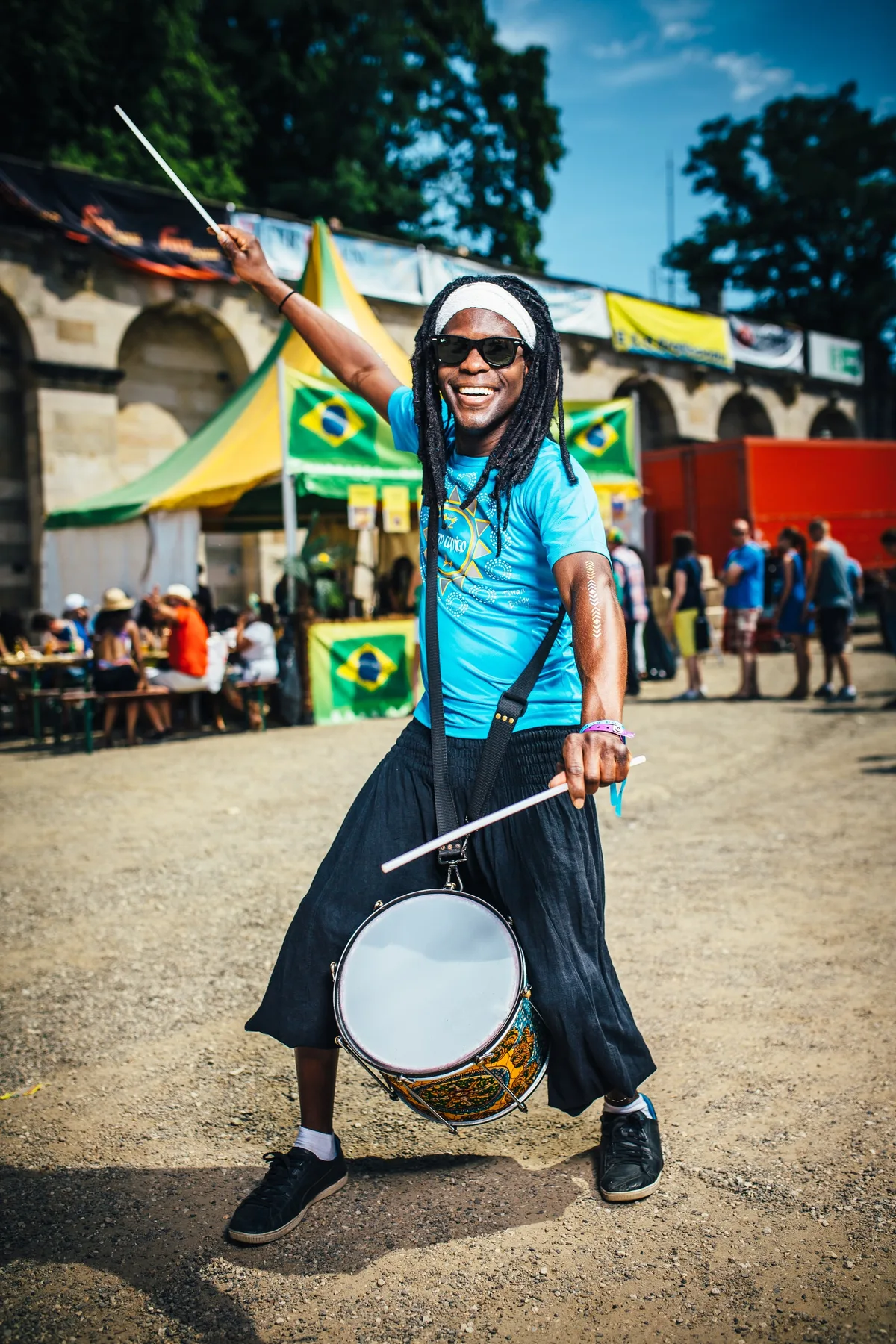 Trommler mit weißem Stirnband und Sonnenbrille hebt lachend einen Stock, hinter ihm Brasilien-Flaggen