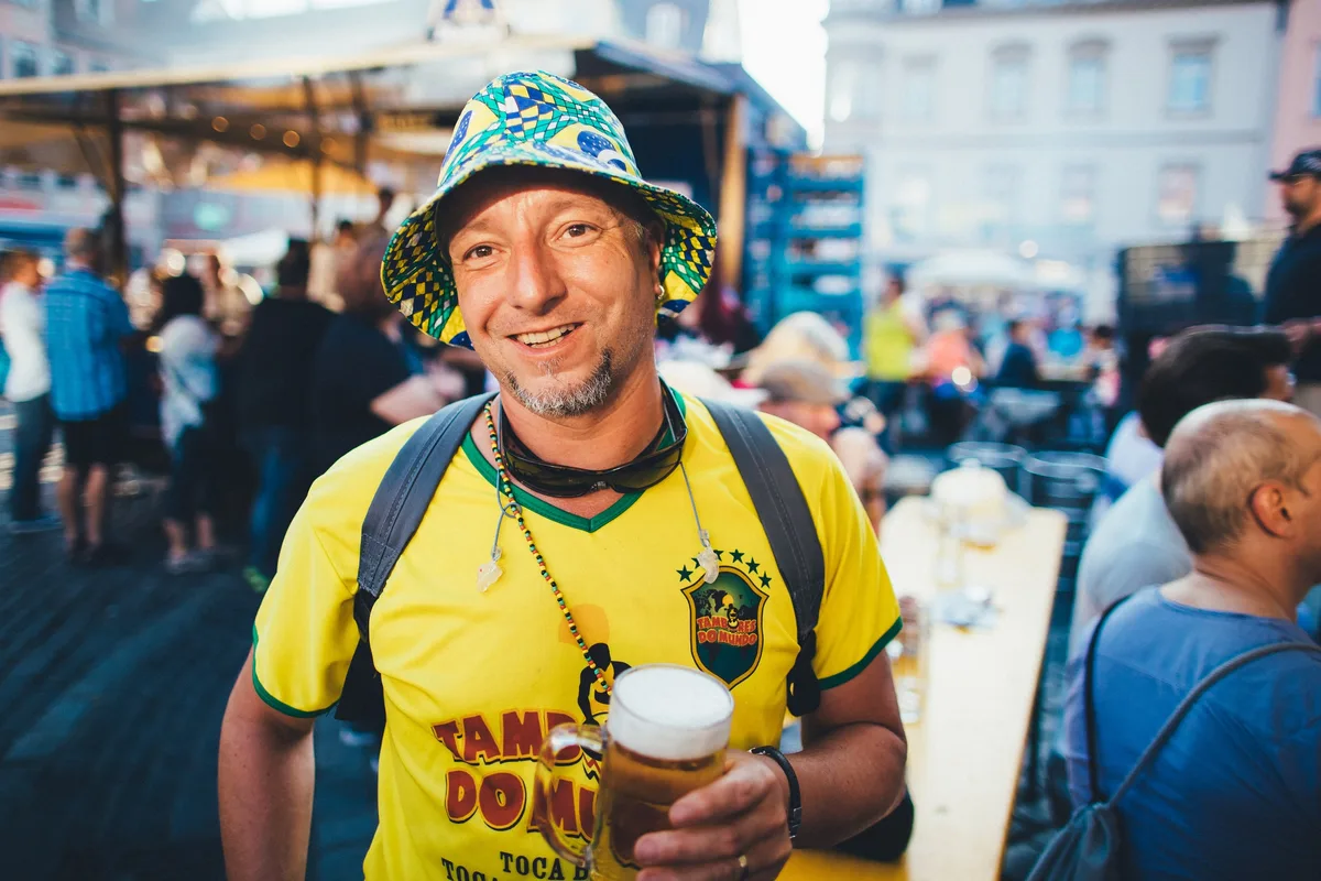 Mann mit buntem Fischerhut und gelbem Brasilien-Trikot hält ein Bierglas, hinter ihm Biertische bei Abendlicht