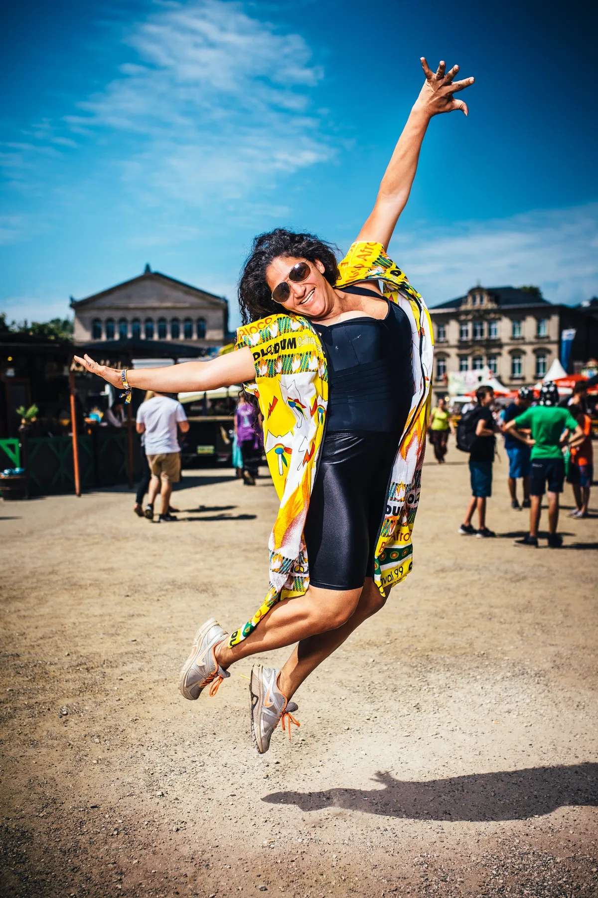 Frau mit Sonnenbrille und gelbem Überwurf springt mit ausgestreckten Armen in die Luft, hinter ihr das Festivalgelände
