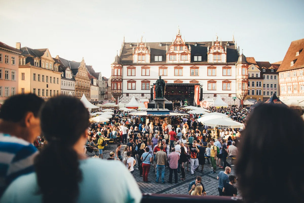 Menschenmenge auf dem Marktplatz mit Bühne und Standbild, davor angeschnittene Köpfe von Zuschauern