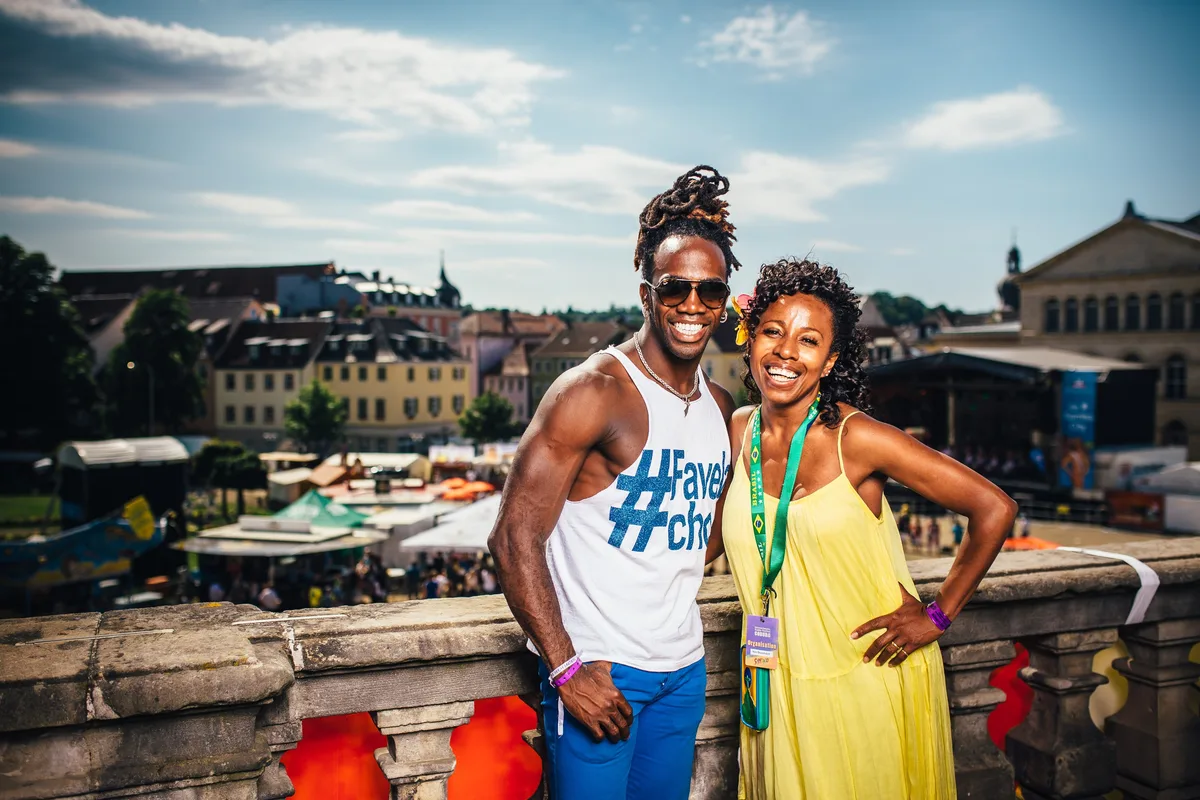 Mann im Trägershirt und Frau im gelben Kleid lehnen lächelnd an einer Steinbalustrade, hinter ihnen das Festivalgelände