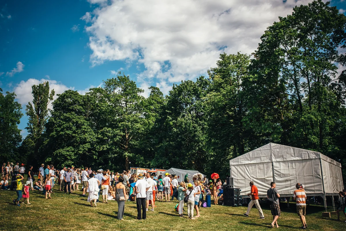 Besucher stehen und sitzen im Park vor einem weißen Zelt und hohen Bäumen, Sommerhimmel mit Wolken