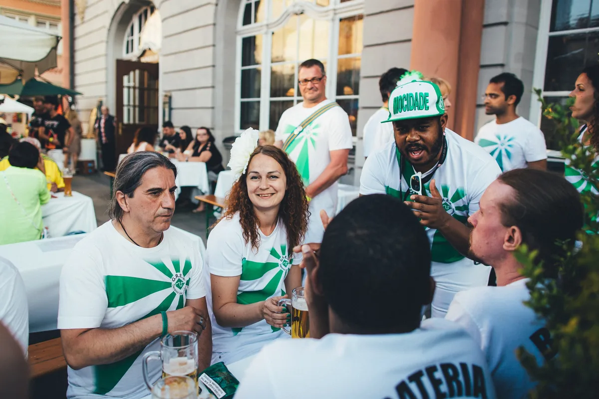 Gruppe in weißen Shirts mit grünem Strahlenmuster sitzt bei Bier im Freien, ein Mann mit grüner Kappe redet mit den Händen