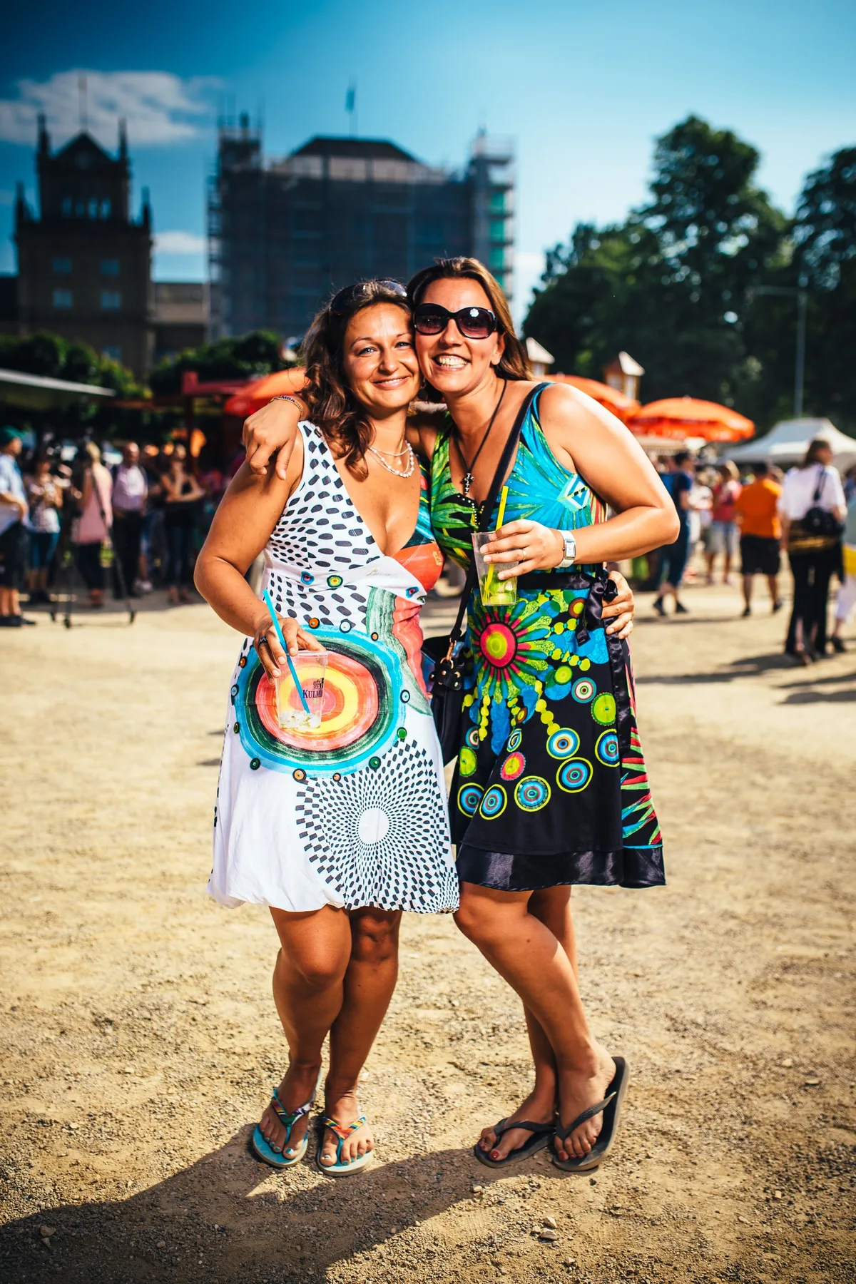 Zwei Frauen in bunt gemusterten Kleidern umarmen sich lächelnd, eine mit Sonnenbrille und Getränk, dahinter ein Gerüstbau