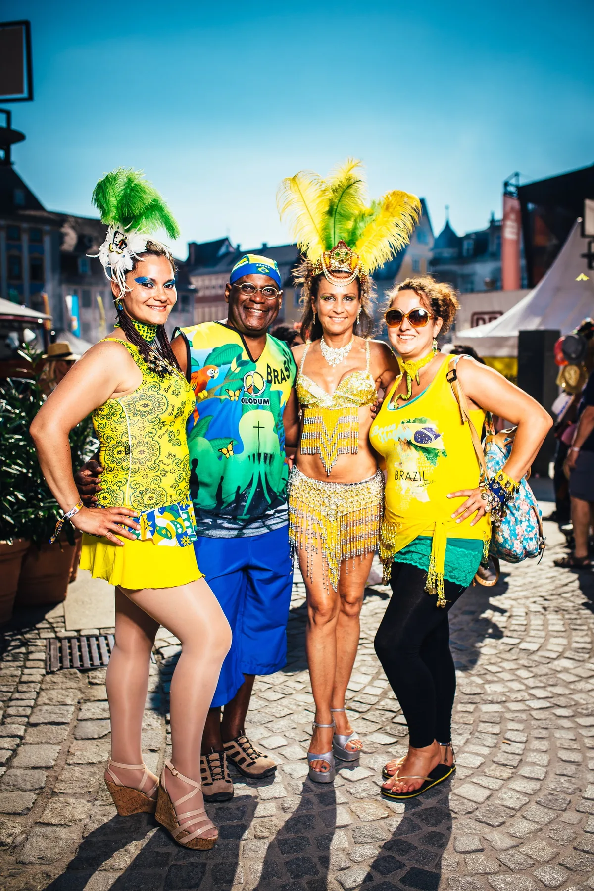 Vier Gäste in Grün und Gelb mit Federkopfschmuck und Brasilien-Shirt stehen Arm in Arm auf Kopfsteinpflaster