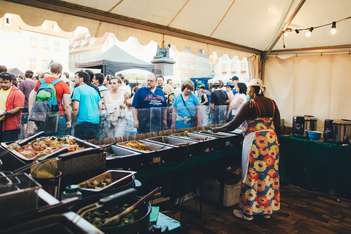 Unter einem Festzelt füllt eine Frau an einer Warmhaltetheke Essen aus, davor eine dichte Menschenschlange