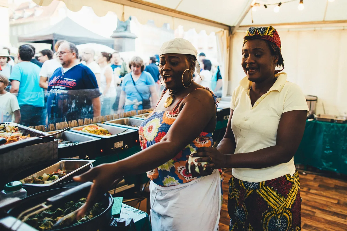 Zwei Frauen in Küchenkleidung bereiten lachend und singend Essen an einer Buffettheke im Zelt zu