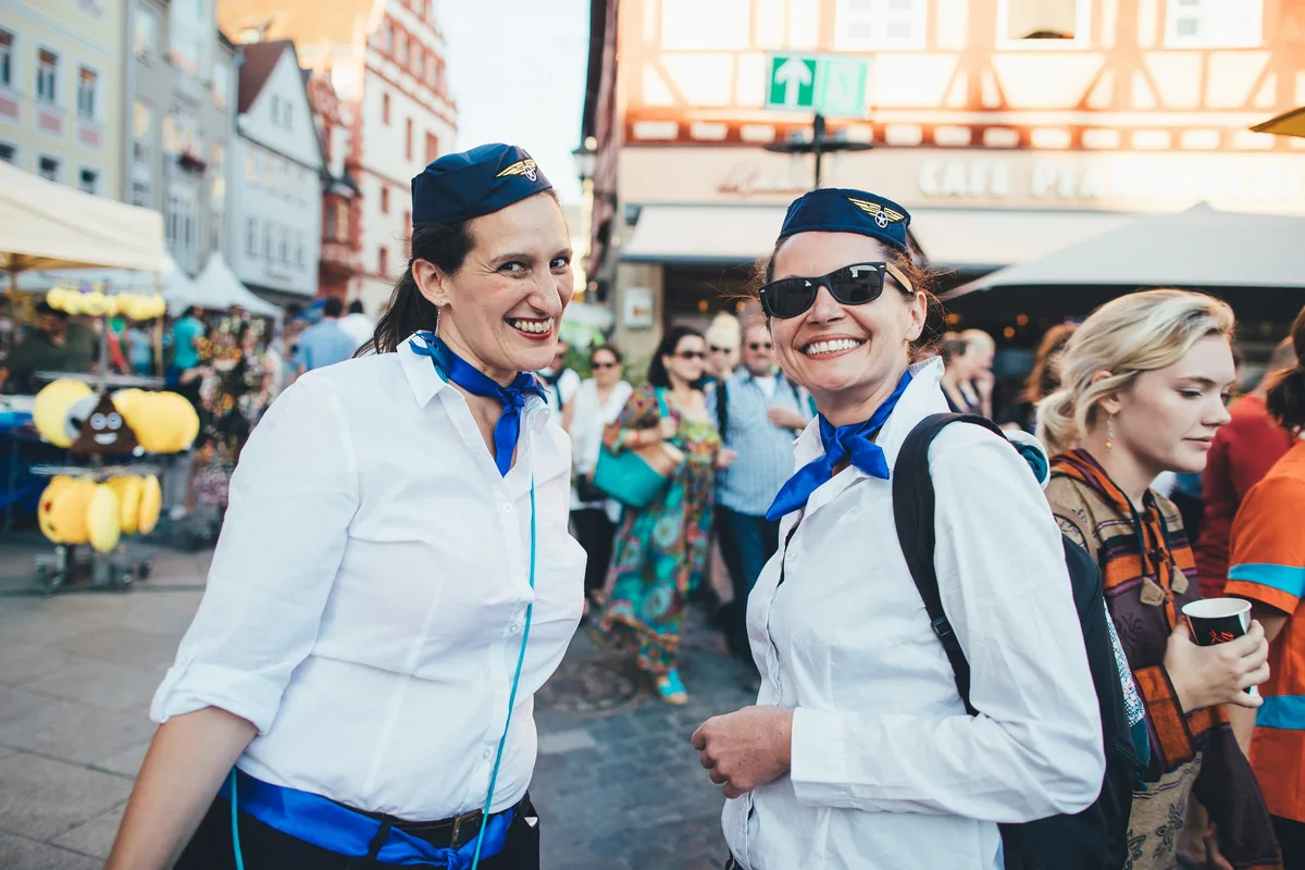Zwei lächelnde Frauen in Fliegerkostümen mit blauen Halstüchern und Schiffchenmützen auf einer Altstadtgasse