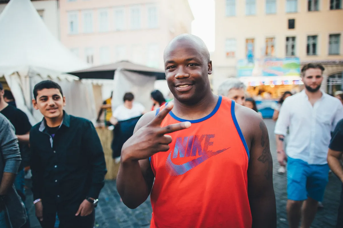 Glatzköpfiger Mann in orangem Nike-Trägershirt zeigt das Peace-Zeichen, hinter ihm ein Jugendlicher und weiße Zelte
