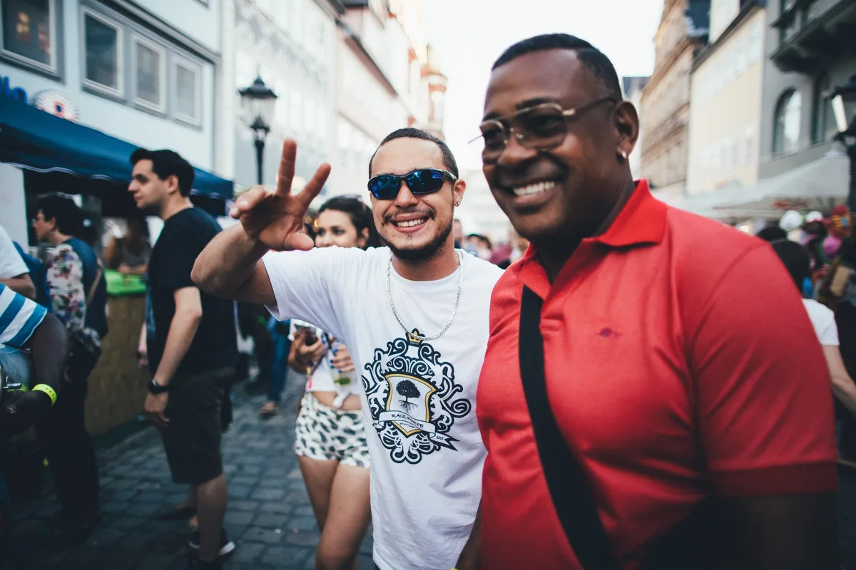 Mann mit blauer Spiegelbrille zeigt das Victory-Zeichen, daneben lacht ein Mann im roten Poloshirt, Gasse im Hintergrund