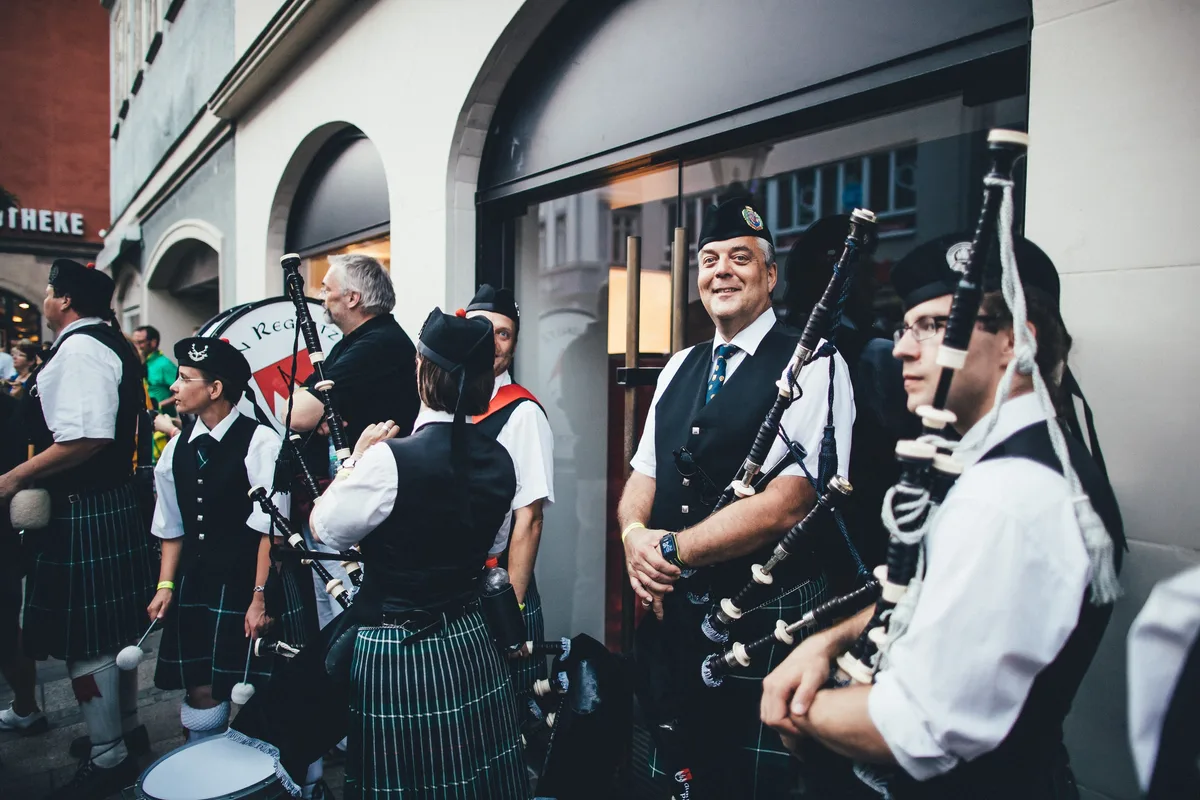 Dudelsackspieler in Kilts und schwarzen Westen stehen vor einem Schaufenster, der Mann in der Mitte lächelt in die Kamera
