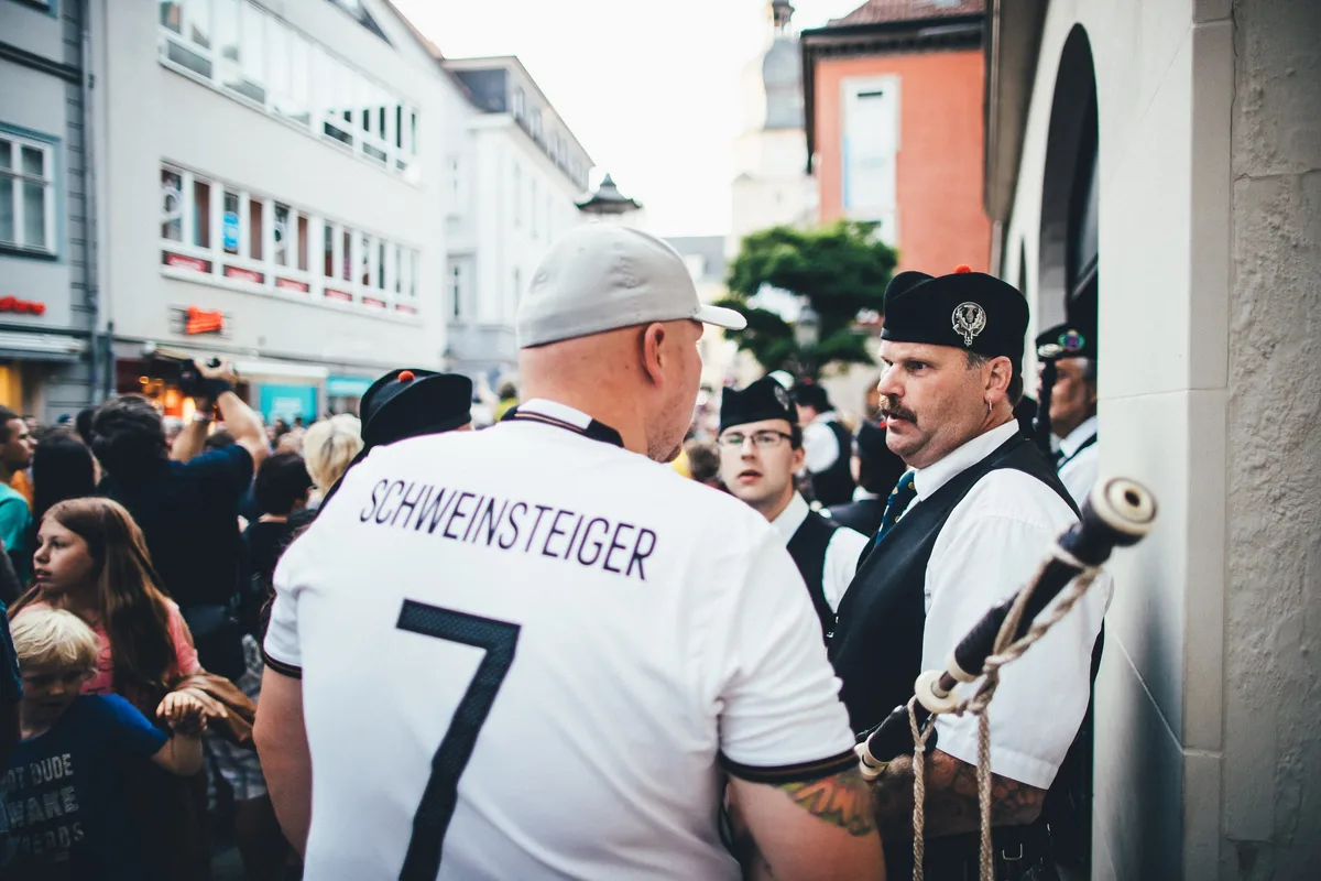 Mann in Deutschland-Trikot mit Schweinsteiger-Aufdruck von hinten im Gespräch mit einem Dudelsackspieler in Kilt-Uniform