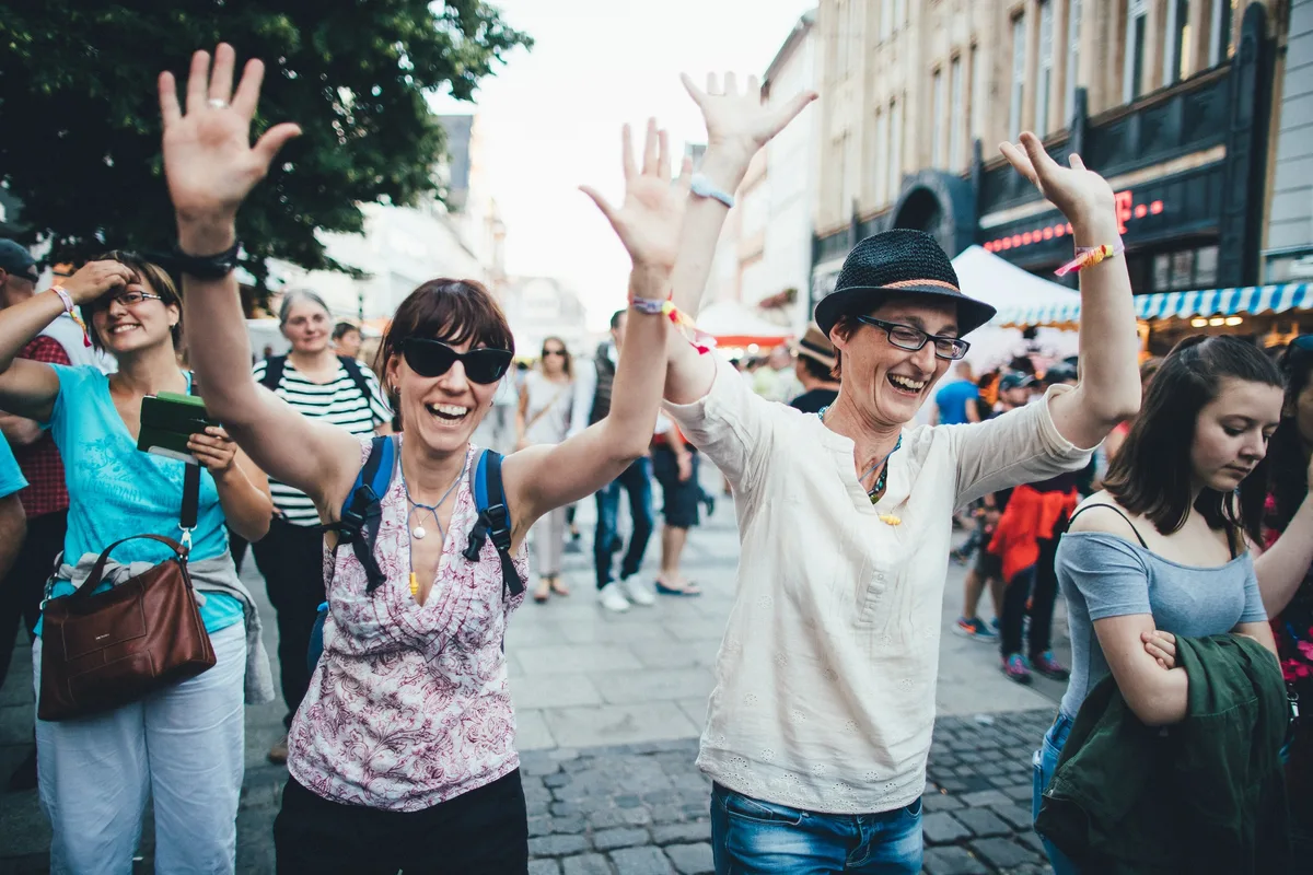 Zwei Frauen mit erhobenen Armen lachen und tanzen auf einer Pflasterstraße unter Menschen