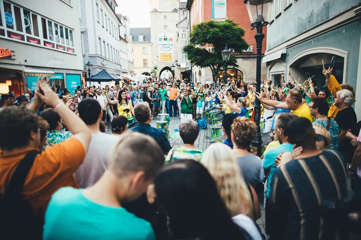 Straßentrommler in Grün spielen auf einer Altstadtgasse, ringsum Zuschauer mit erhobenen Armen, im Hintergrund ein Stadttor