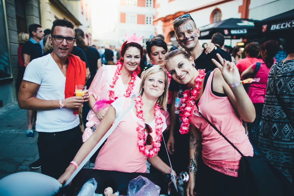 Gruppe in pinkfarbener Kleidung mit rosa Blumenketten lächelt in die Kamera, einer mit Katzen-Gesichtsbemalung und Krönchen