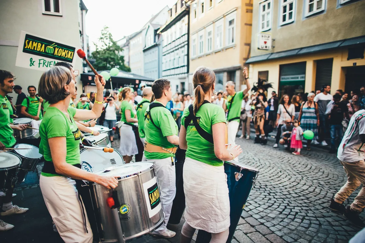 Straßentrommlergruppe in grünen Shirts spielt vor Zuschauern in einer Gasse, links ein Schild mit Gruppennamen