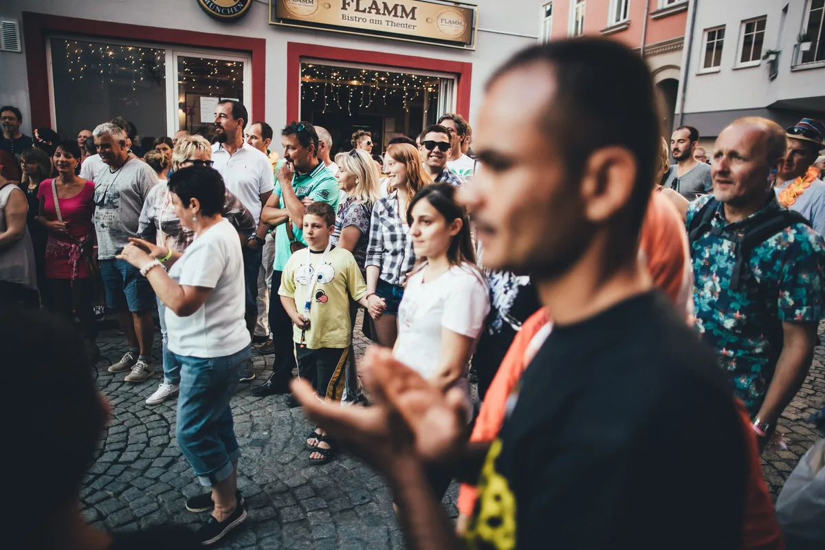 Zuschauer im Gedränge einer Pflastergasse klatschen, im Vordergrund unscharf ein Mann mit angeschnittenem Gesicht