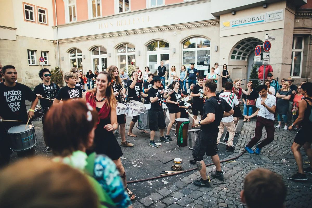 Trommlergruppe in Schwarz spielt auf einer Straße vor Altstadtgebäuden, eine Frau im roten Top lacht in die Kamera