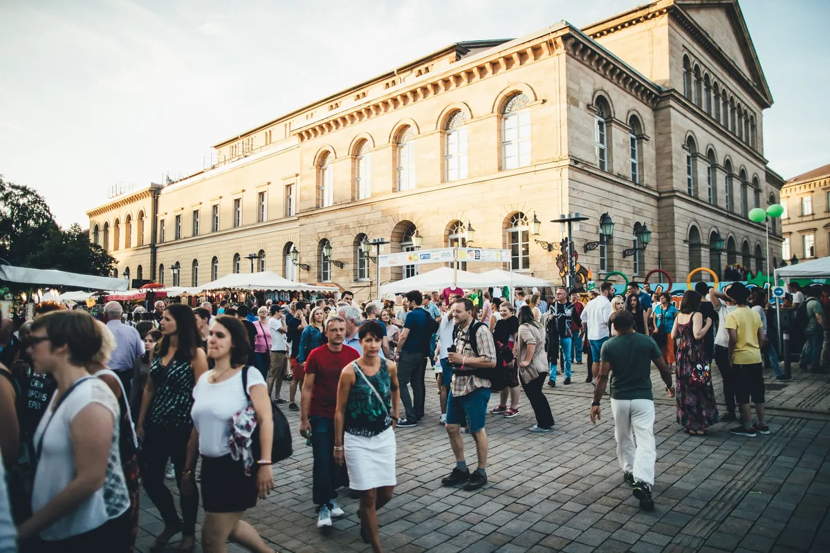 Weite Aufnahme: Besucher strömen über den gepflasterten Platz vor dem klassizistischen Theaterbau, dahinter Stände