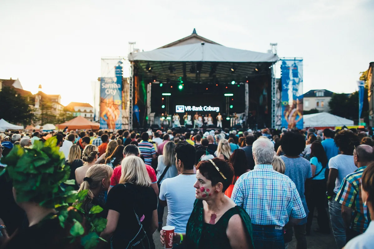 Menge blickt auf die beleuchtete Festivalbühne in der Dämmerung, im Vordergrund Frau mit Blütengesichtsschminke und Becher