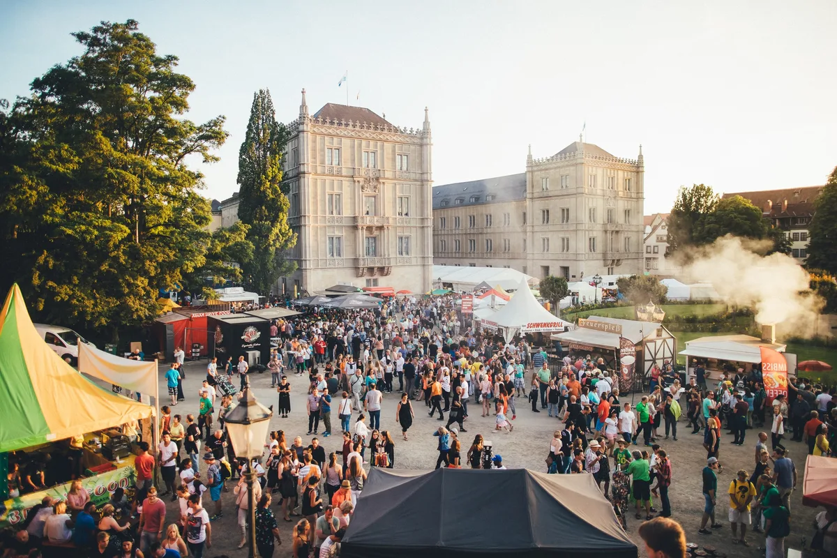 Vogelperspektive auf den Festplatz im Abendlicht, Menschen zwischen Zelten und Ständen vor einem Schloss mit Zinnen