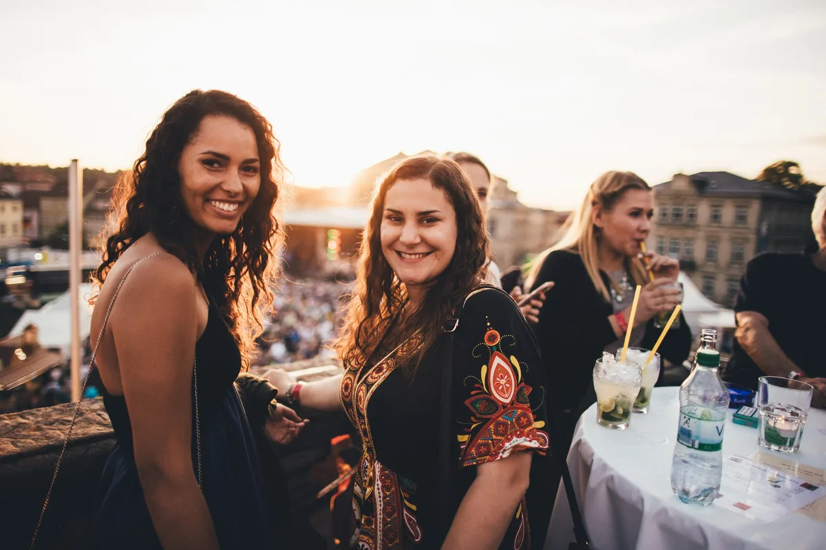 Zwei lächelnde Frauen auf einer Terrasse über dem Festivalgelände, vor ihnen Cocktails mit Strohhalmen auf einem Tisch