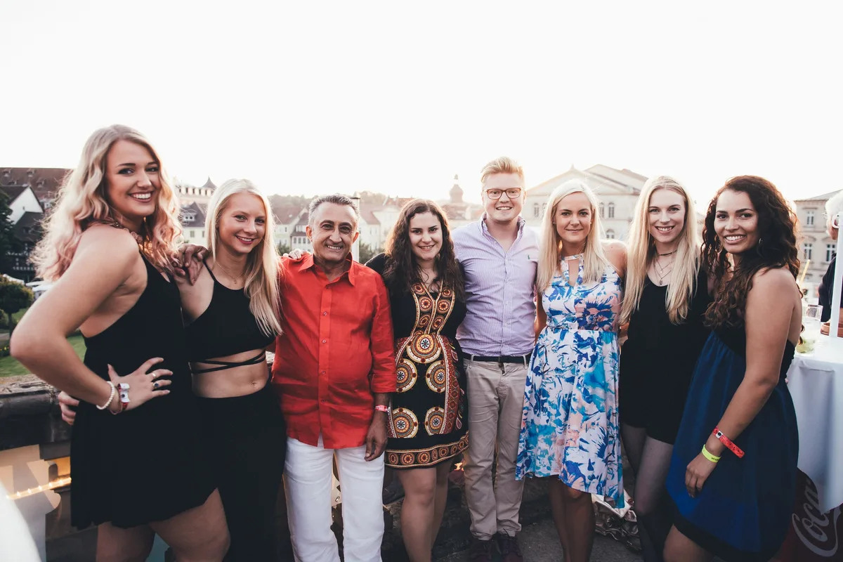 Gruppe aus acht Personen in Sommerkleidung steht nebeneinander auf einer Terrasse und lächelt in die Kamera