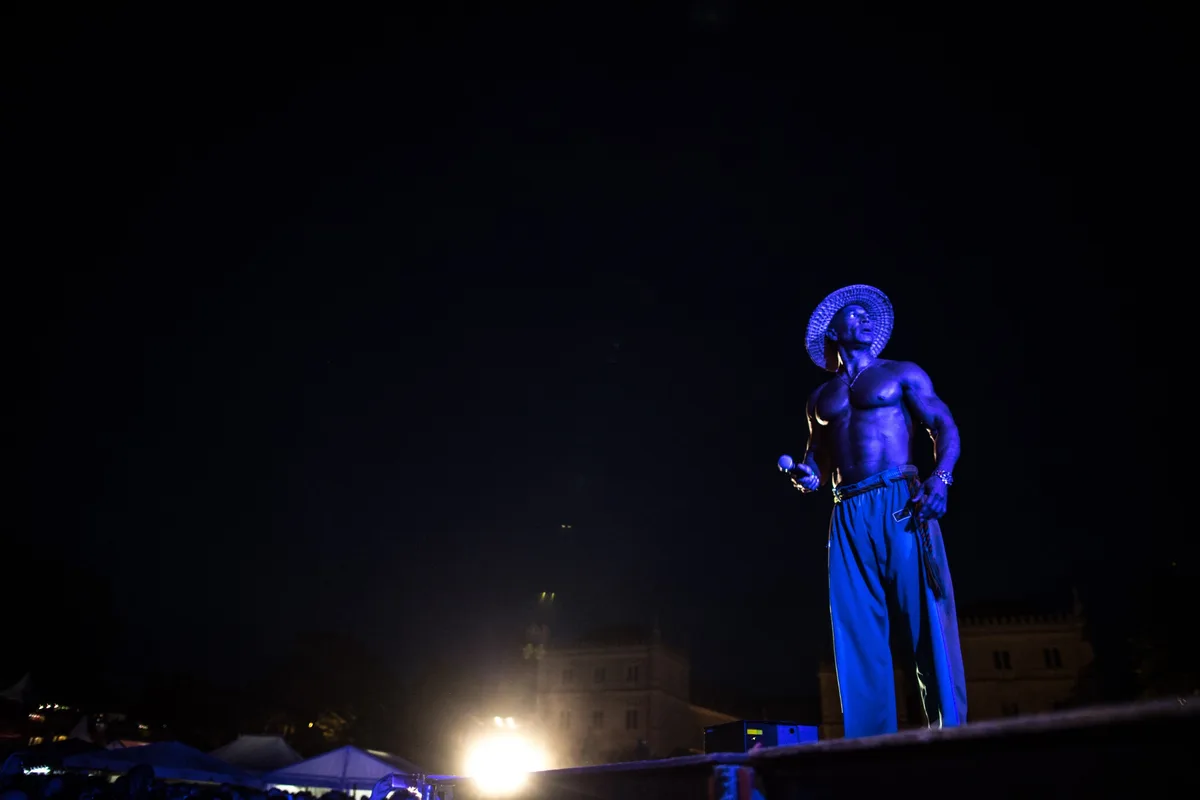Oberkörperfreier Sänger mit Strohhut und Mikrofon steht in blauem Licht auf der Bühne, dunkler Nachthimmel