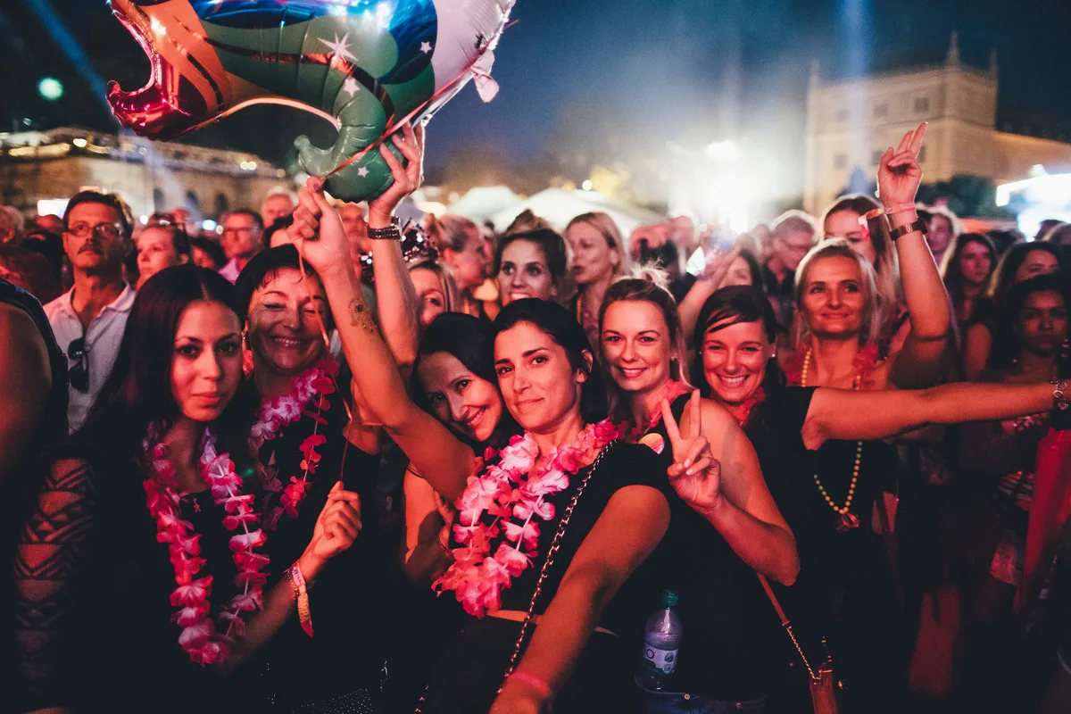 Frauen mit rosa Blumenketten im Publikum bei Nacht, eine zeigt das Peace-Zeichen, über ihnen ein Folienballon