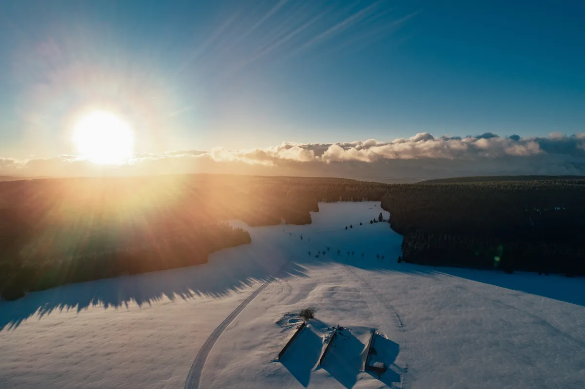 Luftbild einer verschneiten Hochfläche bei tiefstehender Sonne, unten drei kleine Hütten, links und rechts dunkler Wald