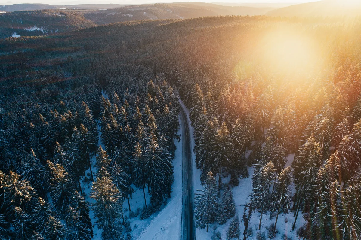 Luftbild einer schmalen Straße zwischen verschneiten Fichten, rechts die Sonne als Lichtfleck über bewaldeten Hügeln