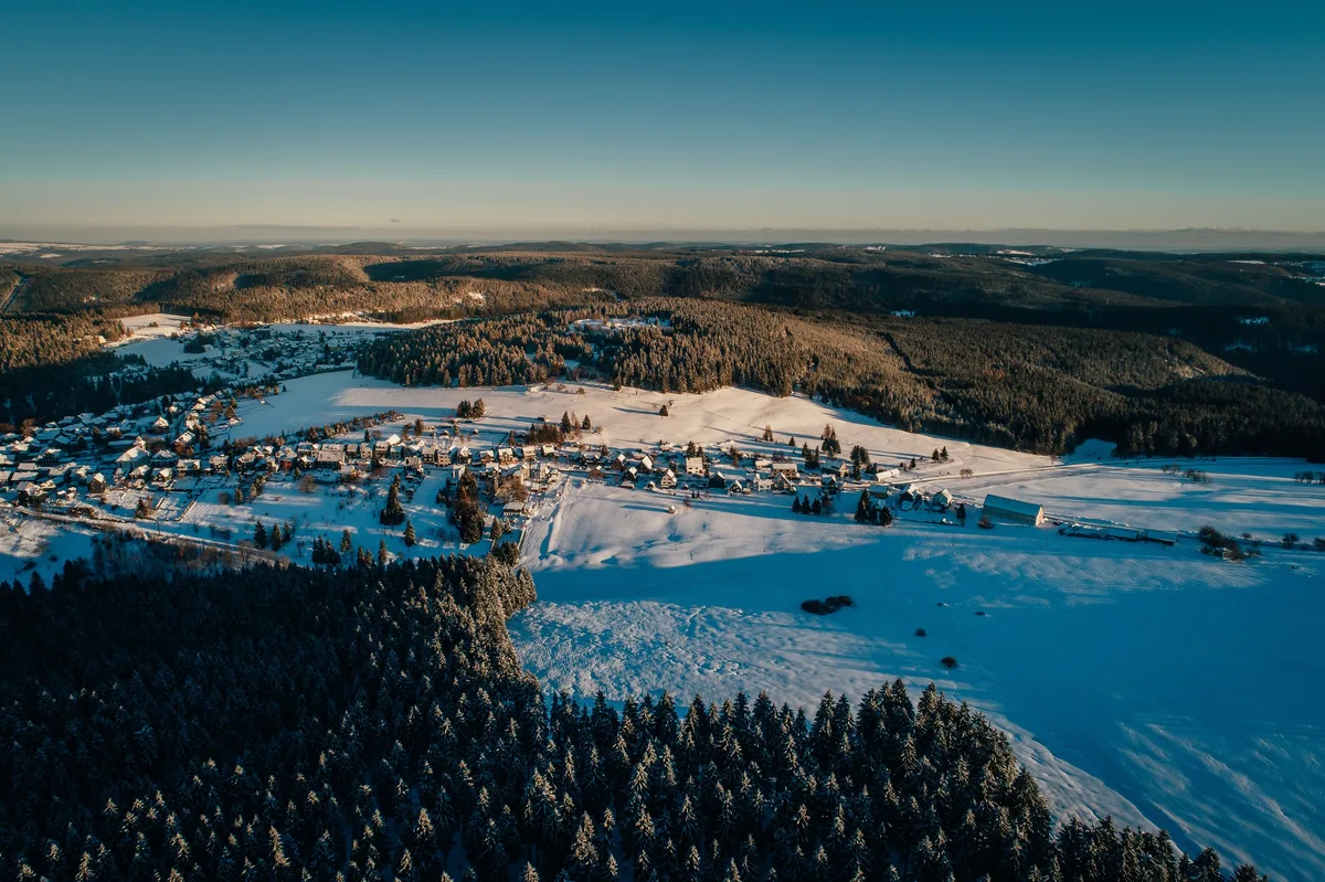 Luftbild eines Dorfes in einer verschneiten Senke, ringsum Wald und weiße Felder, darüber ein blassblauer Himmel
