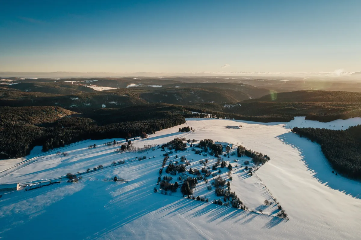Luftbild einer verschneiten Hochebene mit einzelnen Gehöften, lange Baumschatten im Schnee, dahinter Waldhügel im Dunst