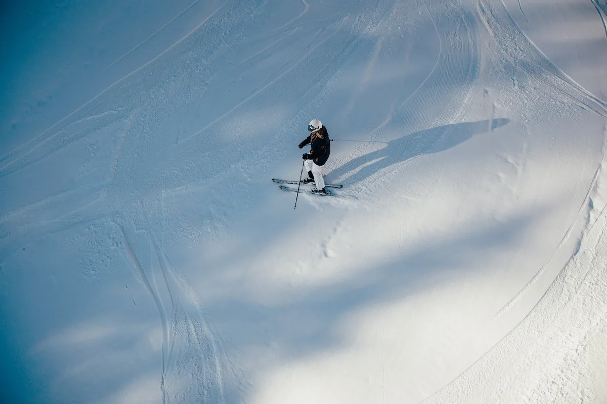 Von oben gesehen fährt eine Skifahrerin mit weißem Helm über einen spurenreichen Hang, ihr Schatten fällt nach rechts