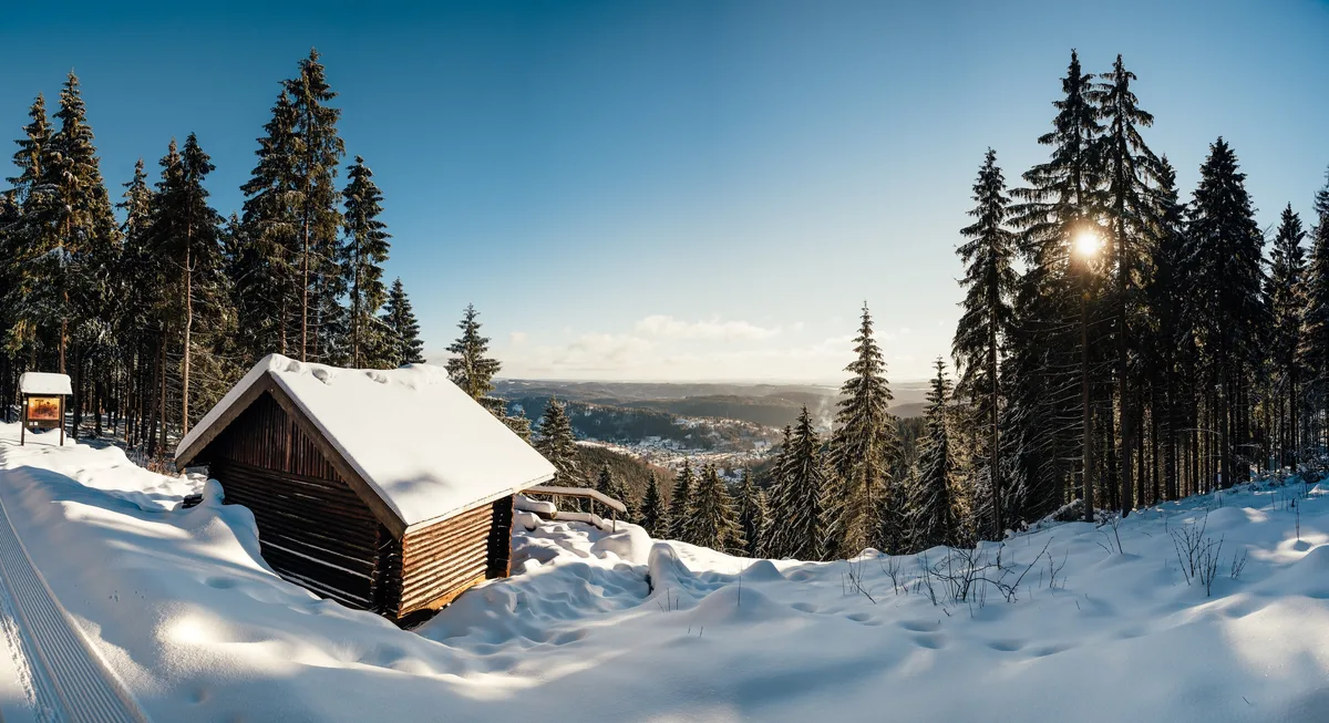 Panorama mit einer kleinen Holzhütte unter Schneedach auf einem Hang, durch die Fichten blickt man in ein Tal mit Ortschaft